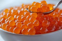 Fresh red caviar in white ceramic bowl with silver spoon, macro shot showing detailed texture of salmon roe pearls with natural glossy shine.