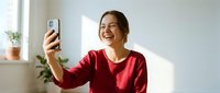 Happy young Caucasian woman in red sweater taking selfie with smartphone at home, laughing and smiling while video calling or photographing herself indoors.