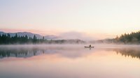 Serene lake at sunrise with mist rising from calm water, lone fisherman in boat, forested shoreline, peaceful morning atmosphere, soft pink and purple sky reflecting on glassy surface.