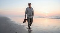 Middle-aged Caucasian man in casual business attire walking barefoot on wet sand beach at sunset, carrying shoes in hand, relaxed lifestyle and freedom concept.