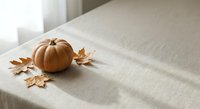 Orange pumpkin with autumn maple leaves on beige linen tablecloth in soft natural light creating cozy fall seasonal home decor atmosphere.