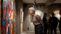 Elegant senior woman with silver hair admiring colorful abstract artwork in contemporary art gallery with exposed brick walls and track lighting.