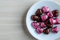 Heart-shaped chocolate candies wrapped in pink and brown foil arranged on white ceramic plate against light wooden surface, romantic holiday concept.