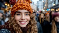 Young Caucasian woman with curly hair wearing orange knitted winter hat and sweater smiling at camera on busy city street with bokeh lights and crowd in background.