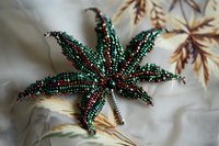 Handcrafted beaded marijuana leaf brooch with colorful green and red seed beads on fabric background with autumn leaf pattern.