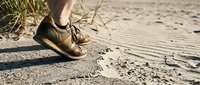 Unrecognizable person wearing brown shoes standing on sandy beach with dry grass and footprints visible in sand during daytime.