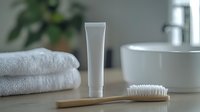 White cosmetic tube with wooden toothbrush and folded towels on bathroom counter, minimalist spa composition against blurred modern interior background.