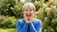 Mature woman with short blonde hair, Caucasian ethnicity, wearing blue cardigan, expressing joy and surprise with hands on face in garden setting with blooming flowers.