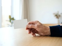 Senior adult hand holding blank white business card on wooden table with natural window light, suitable for corporate identity, networking, and professional branding mockups.