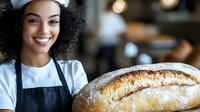 Young African American female baker with curly hair and bright smile holding fresh artisanal bread loaf in bakery. Professional food service worker showcasing homemade baked goods.
