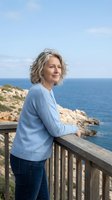 Mature Caucasian woman with gray hair enjoying peaceful moment on coastal balcony overlooking blue ocean and rocky cliffs on sunny day.