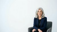 Mature woman with gray hair wearing dark blazer seated in gray chair against white background, professional portrait for corporate business use.