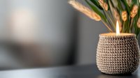 Cozy knitted candle holder with burning candle and dried wheat stalks creating warm atmospheric lighting against blurred background. Hygge home decor concept.