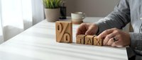 Businessperson arranging wooden blocks displaying percentage and tax symbols on white desk, representing financial planning, accounting, and tax calculation concepts for business and finance.