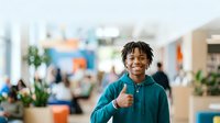 Young male student with dark skin tone wearing teal hoodie giving thumbs up in modern school hallway with blurred classmates and colorful educational environment in background.