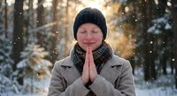 Young Caucasian woman in winter coat and knit hat with hands in prayer position during snowfall in forest. Peaceful meditation and gratitude concept.