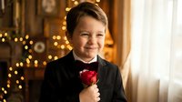 Young boy in formal black suit holding red rose with warm Christmas lights creating romantic holiday atmosphere for special occasions.