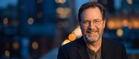 Smiling mature businessman with glasses and beard in dark suit against blurred city lights background during evening hours for corporate portraits.