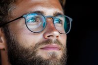 Close up portrait of young Caucasian man with beard wearing round glasses, illuminated by blue light reflection, looking up with thoughtful expression.