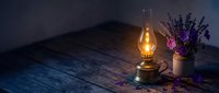 Vintage oil lamp glowing warmly on rustic wooden table with dried lavender flowers in ceramic vase creating cozy atmospheric lighting for home decor.