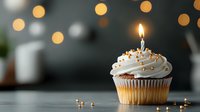 Single vanilla cupcake with white frosting, lit birthday candle and gold sprinkles on dark background with festive bokeh lights, celebration concept.