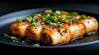 Grilled squid tubes garnished with sesame seeds, green onions and chili flakes served on black plate, close up view with selective focus and dark background.