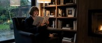Young woman reading book in cozy armchair by fireplace with wooden bookshelves in warm living room interior for relaxation and leisure concepts.