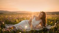 Young pregnant woman in white dress relaxing on blanket in wildflower meadow during golden hour sunset with warm sunlight and peaceful atmosphere.