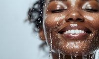 Close-up of African American woman with water splashing on face, eyes closed, showing bright smile and glowing skin during skincare routine.