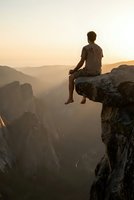 Young man sitting on cliff edge at sunset overlooking mountain valley landscape during golden hour meditation moment for mindfulness and adventure concepts.
