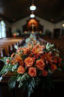 Orange roses and lilies arranged in elegant funeral casket spray with lush greenery, photographed in church sanctuary with soft lighting and wooden pews.
