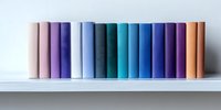 Row of colorful hardcover books standing on white shelf against light wall, arranged in gradient spectrum from pink through blue to orange tones.