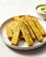 Crispy golden zucchini sticks with herbs served on white plate with creamy avocado dip, perfect for healthy appetizers or party snacks.