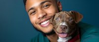 Happy young African American man smiling while holding adorable brown and white pit bull puppy against teal background for pet adoption campaigns.