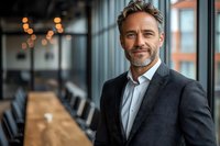 Confident mature businessman with gray hair and beard wearing dark suit jacket smiles in modern office setting with bokeh lights and conference room background.
