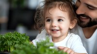 Joyful toddler girl with curly hair smiling while exploring fresh parsley with bearded father in kitchen, healthy eating and family bonding concept.
