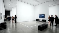 Contemporary art gallery interior with polished concrete floor, white walls, and minimalist black seating. Multiple visitors explore modern exhibition space with digital display and natural lighting for cultural venues.