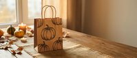 Autumn pumpkin pattern kraft paper shopping bag on wooden table with fall leaves and decorations for seasonal retail packaging and harvest themed gifts.