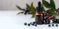 Brown glass dropper bottle with fresh elderberries and green leaves scattered on white surface, natural wellness concept for aromatherapy and herbal medicine.