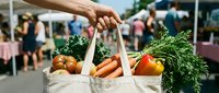 Farmer's hand holding canvas tote bag filled with fresh organic vegetables at outdoor market, featuring carrots, tomatoes, and leafy greens for sustainable shopping.