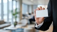 Businessman in formal suit holding blank white business card in modern office environment with colleagues working in background.