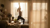 Young woman practicing yoga warrior pose in sunlit living room with warm golden light streaming through curtains creating peaceful morning wellness atmosphere.