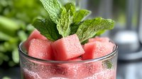 Fresh watermelon cubes garnished with mint leaves in glass, refreshing summer drink or dessert concept with condensation droplets on transparent surface.
