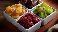 Colorful assortment of dried fruits in white ceramic bowls including cranberries, apricots, dates and sliced kiwi on wooden table, healthy snack variety.