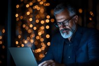 Mature Caucasian businessman with gray beard and glasses working on laptop at night, illuminated by warm bokeh lights creating cozy atmosphere.