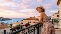 Young Caucasian woman in floral dress and sun hat enjoying sunset view from Mediterranean balcony overlooking coastal town and sea during summer vacation.