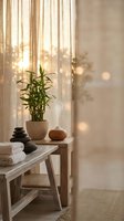 Serene spa interior with bamboo plant, stacked stones and towels on wooden bench near window with warm sunlight filtering through curtains creating peaceful atmosphere.