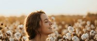 Young woman with curly hair enjoying peaceful moment in cotton field during golden hour sunset, eyes closed in serene expression for wellness and nature concepts.