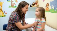 Female pediatrician examining young Caucasian girl with stethoscope in colorful children's medical office with animal wall decorations during routine checkup.