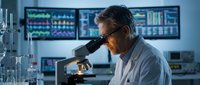 Middle-aged Caucasian male scientist examining samples through microscope in modern laboratory with digital monitors displaying data analysis results.